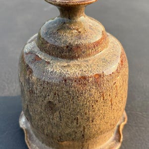 May include: A ceramic oil lamp with a mottled brown and tan glaze. The lamp features a rounded body, a narrow neck, and a flared, scalloped base. A small, circular ceramic insert sits at the top opening, holding a single, short, dark fabric wick.