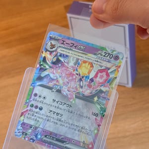 May include: A Japanese Espeon ex trading card featuring a crystalline Terastal design with pink, blue, and purple accents. The card displays 270 HP, the name "エーフィex", and various game text in Japanese, all encased in a clear protective sleeve.
