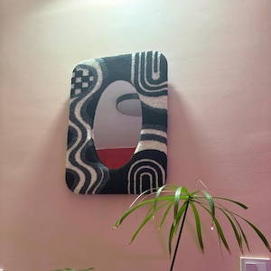 May include: A rectangular wall mirror featuring a tufted, textured frame in a black, white, and gray color palette. The frame displays a pattern of wavy lines, a checkered square in the top left corner, and rounded arch shapes along the sides.