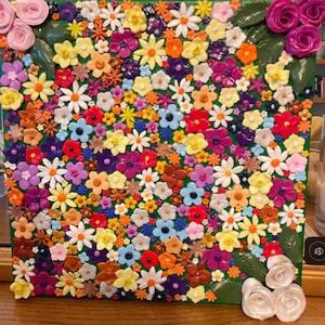 May include: A square wall art piece featuring a dense, colourful arrangement of small, three-dimensional sculpted flowers in shades of pink, yellow, orange, purple, blue, and white, accented with green leaves and clusters of larger roses.