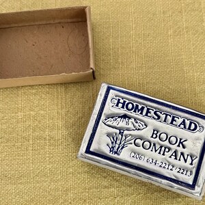 May include: A rectangular silver-toned metal box with a blue border and the embossed text HOMESTEAD BOOK COMPANY, a mushroom illustration, and the phone numbers (206) 634-2212/2213, accompanied by an empty, open brown cardboard matchbox tray.