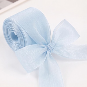 Pode incluir: Um rolo de fita azul-clara com uma textura transparente e um brilho subtil. A fita está enrolada num cilindro apertado, com uma parte do tecido estendida para fora e atada num laço decorativo simples na parte da frente.