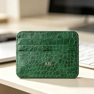 May include: A rectangular dark green card holder featuring a textured crocodile skin pattern. The front displays two horizontal card slots and the gold-coloured embossed initials AB centred on the lower portion of the accessory.