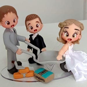 Pode incluir: Um conjunto de três figuras estilo chibi sobre uma base transparente. Uma figura de terno cinza empurra um carrinho de mão cinza contendo uma figura de vestido branco. Uma segunda figura de terno preto está próxima. Inclui tijolos laranja e um saco com a etiqueta CIMENTO.