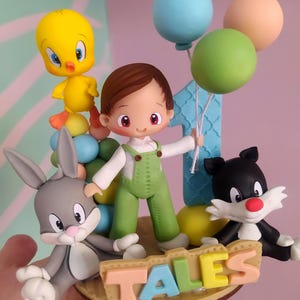 Puede incluir: Un adorno para pastel con una figura central en overol verde, flanqueada por un pájaro amarillo, un conejo gris y un gato blanco y negro. Un número uno azul se alza detrás, con tres globos y un letrero con la palabra TALES.