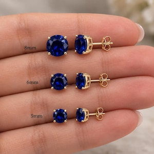 May include: Three pairs of round, deep blue stud earrings set in gold-toned metal prongs. The earrings are displayed in three distinct sizes labeled as 8mm, 6mm, and 5mm, each featuring a faceted gemstone and a standard post-back closure mechanism.