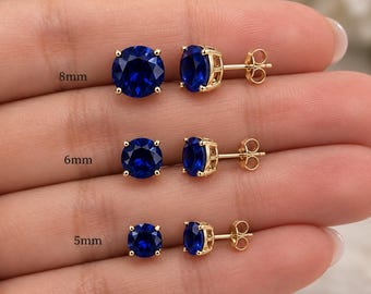 May include: Three pairs of round, deep blue stud earrings set in gold-toned metal prongs. The earrings are displayed in three distinct sizes labelled as 8mm, 6mm, and 5mm, each featuring a faceted gemstone and a standard post-back closure mechanism.