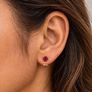 May include: A round, faceted red gemstone stud earring set in a gold-toned metal prong setting. The earring is positioned on an earlobe, with a small graphic indicator below it showing a diameter measurement of 5mm for the stone.