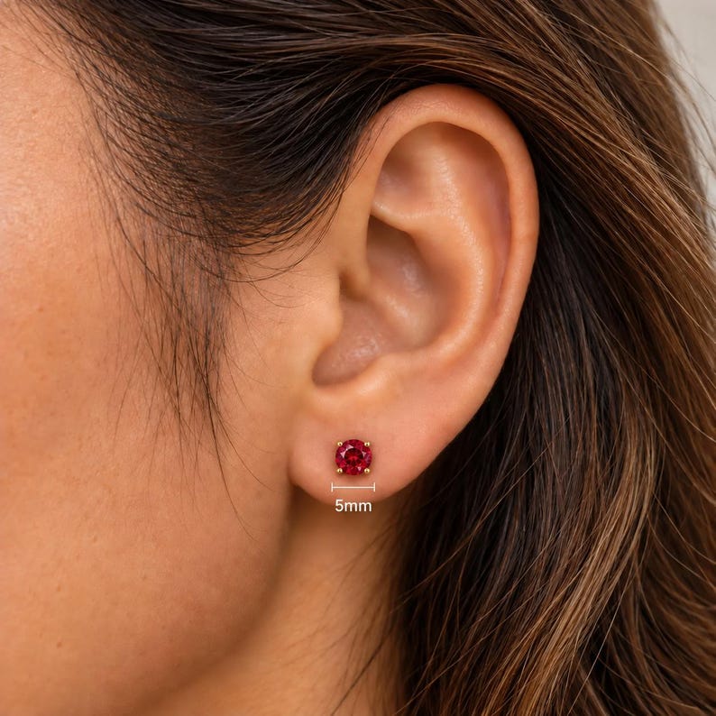 May include: A round, faceted red gemstone stud earring set in a gold-toned metal prong setting. The earring is positioned on an earlobe, with a small graphic indicator below it showing a diameter measurement of 5mm for the stone.