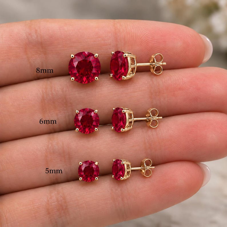 May include: Three pairs of round-cut red gemstone stud earrings set in gold-toned metal. The pairs are labelled with sizes 8mm, 6mm, and 5mm. Each earring features a four-prong setting and a standard post back, showcasing a vibrant red colour.