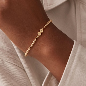 Op de afbeelding: Een goudkleurige kralenarmband bestaande uit een aaneengesloten rij kleine, uniforme en gepolijste kralen. Het sieraad is voorzien van twee kleine, ronde bedels die elk zijn gegraveerd met een enkele letter, namelijk de letters T en J.