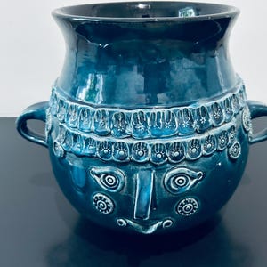 Puede incluir: Un recipiente de cerámica en color verde azulado profundo con dos asas laterales y un borde ancho y acampanado. El exterior presenta un patrón en relieve de rasgos faciales humanos estilizados, incluyendo ojos almendrados y una nariz central.