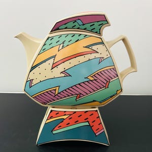 Könnte beinhalten: Eine cremefarbene Teekanne mit geometrischer, kantiger Form und Deckel. Der Körper und der Sockel zeigen ein lebhaftes, mehrfarbiges Muster aus türkisfarbenen, rosa, gelben und grünen Zickzackformen, akzentuiert durch schwarze Striche und Punkte.