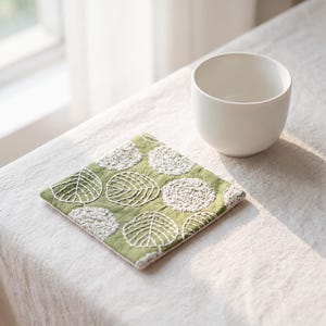 May include: A square fabric coaster featuring a green background with white embroidered circular patterns and leaf motifs, accompanied by a plain white ceramic cup. The coaster has a flat, quilted texture with visible white thread stitching.