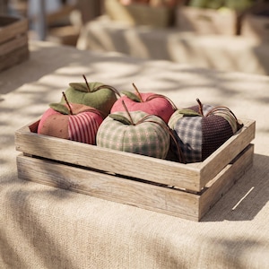 Peut inclure: Une caisse en bois rectangulaire contenant cinq pommes en tissu. Chaque pomme présente un motif textile unique, incluant des rayures rouges, des carreaux verts et des motifs sombres. Chaque pomme est surmontée d'une petite feuille en tissu vert.