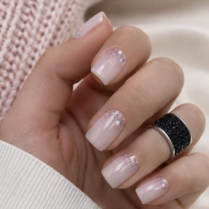 May include: A wide silver-toned ring featuring a textured black surface worn on a finger with square-shaped nails. The nails are painted in a pale pink gradient finish with silver glitter accents concentrated near the cuticle area of each nail.