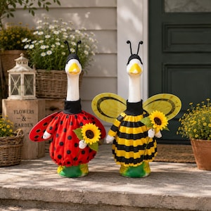 May include: Two goose figurines dressed in costumes. One wears a red dress with black polka dots and black antennae, holding a sunflower. The other wears a yellow and black striped dress with yellow wings and black antennae, holding a sunflower.