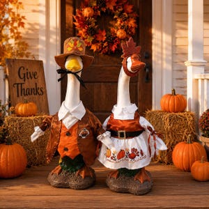 May include: Two goose figurines dressed in autumn-themed clothing. The left goose wears a brown hat and vest with a turkey pin. The right goose wears a leaf headband and a white dress with pumpkin and turkey embroidery. Both stand on stone bases.