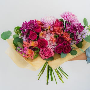 Könnte beinhalten: Ein Blumenstrauß mit dunkelmagentafarbenen Rosen, hellrosa Chrysanthemen, orangefarbenen Inkalilien und violetten Levkojen, akzentuiert durch grüne Eukalyptusblätter und eingewickelt in durchscheinendes beiges Papier mit sichtbaren grünen Stielen.