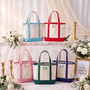 May include: Five canvas tote bags in off-white with colored trim and handles in light blue, red, pink, green, and dark blue. Each bag features a front pocket with embroidered initials: HBT, ST, GS, CD, and HE in matching thread colors.