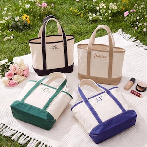 May include: Four canvas tote bags with contrasting colored bases and handles. One bag has a black trim and bow graphic, one is tan with the name Jessica, one is green with a monogram, and one is blue with the initials AF embroidered on the front.