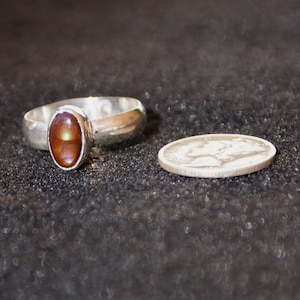 Op de afbeelding: Een zilveren ring met een bruine ovale edelsteen in het midden. De ring ligt op een donkergrijs oppervlak.