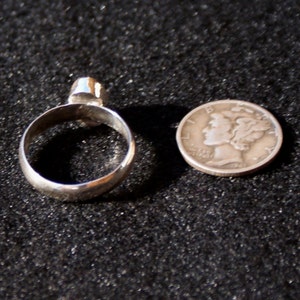 Op de afbeelding: Een zilveren ring met een heldere steen in het midden. De ring wordt getoond naast een Amerikaanse dime ter vergelijking van de grootte.