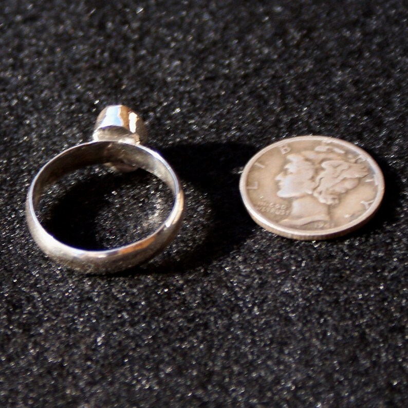 Op de afbeelding: Een zilveren ring met een heldere steen in het midden. De ring wordt getoond naast een Amerikaanse dime ter vergelijking van de grootte.