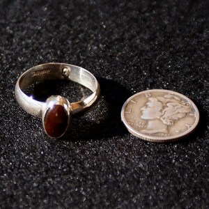 Op de afbeelding: Een zilveren ring met een donkerrode ovale edelsteen. De ring ligt naast een zilveren Amerikaanse dime met het profiel van een vrouwenhoofd en de tekst "In God We Trust" en "1941".