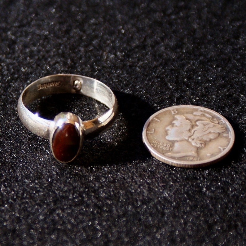 Op de afbeelding: Een zilveren ring met een donkerrode ovale edelsteen. De ring ligt naast een zilveren Amerikaanse dime met het profiel van een vrouwenhoofd en de tekst "In God We Trust" en "1941".