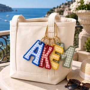 May include: A cream-colored canvas tote bag featuring a gold-toned carabiner clip attached to the handle. Hanging from the clip are four fabric letter charms in a gingham pattern, colored blue, red, yellow, and sage green, spelling out the word AKEM.