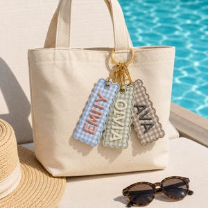 May include: A cream-colored canvas tote bag featuring three rectangular, scalloped-edge fabric keychains attached to a gold-toned ring. The keychains display the names EMILY in peach, OLIVIA in white, and AVA in dark gray on gingham patterns.