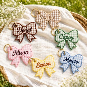 May include: Six embroidered bow-shaped keychains featuring a gingham pattern in brown, green, pink, yellow, and blue. Each keychain includes a gold-tone metal ring and is personalized with the names Evelyn, Donal, Cappy, Misan, Semon, and Mewto.