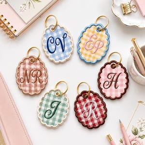 May include: Six oval-shaped keychains with scalloped edges, each featuring a gingham pattern in different colors including blue, yellow, brown, green, and red. Each keychain has a gold-toned metal ring and is embroidered with unique initials.