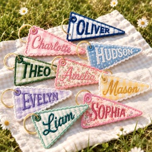 May include: A collection of nine triangular fabric keychains featuring embroidered names including Oliver, Charlotte, Hudson, Theo, Amelia, Mason, Evelyn, Liam, and Sophia. Each keychain has a gold metal ring and scalloped white trim edges.