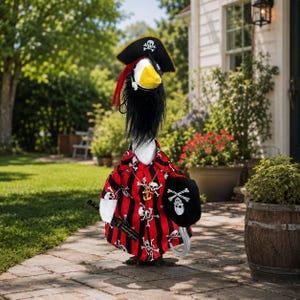 Puede incluir: Una figura decorativa de ganso disfrazada de pirata, con una camisa de rayas negras y rojas con patrones de calaveras y tibias cruzadas, un sombrero de pirata negro con un emblema de calavera, un collar dorado, un parche negro y un gancho plateado.