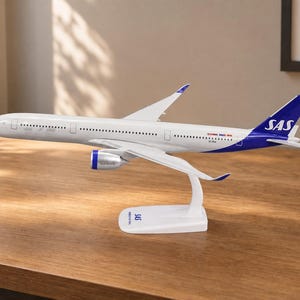 以下が含まれることがあります： 青い尾翼にSASのロゴが入った白い飛行機のスケールモデル。機体は白い胴体に小さな窓の列があり、青いエンジンカウルを備えています。SASのロゴが印字された白い曲線状のスタンドに取り付けられています。