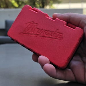 May include: A rectangular red plastic storage case with a textured surface and the Milwaukee brand logo embossed on the front. The case features a hinged lid design with a black base visible along the side edges, providing a compact container.