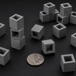 May include: A collection of small, hollow, grey concrete cubes arranged in various positions, including a vertical stack of three. A Canadian 25-cent coin is placed among the cubes to provide a sense of scale for these miniature square objects.