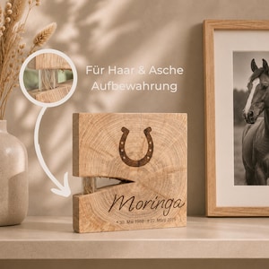 Könnte beinhalten: Ein quadratisches Gedenkobjekt aus Holz mit eingraviertem Hufeisen, dem Namen Moringa sowie den Daten 30. Mai 1988 und 22. März 2019. Ein kleines Glasröhrchen ist in einen horizontalen Schlitz eingelassen, zur Aufbewahrung von Haar und Asche.
