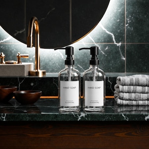 May include: Two clear glass soap dispensers with matte black pump mechanisms. Each bottle features a white rectangular label with the text "HAND SOAP" in black, followed by a horizontal line and the word "ESSENTIAL" printed below it.