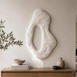 May include: A large, off-white, organic-shaped wall sculpture with a textured, matte surface and a central open void. The piece features an irregular, asymmetrical silhouette with a rough, plaster-like finish, mounted centrally on a plain wall.