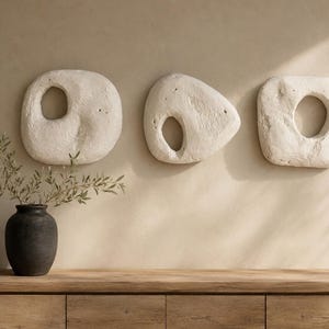 May include: Three off-white, textured, organic-shaped wall sculptures with central cutouts, arranged horizontally. A dark charcoal-colored vase containing a leafy green branch sits to the left on a light-toned wooden console table.