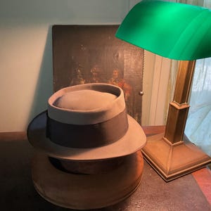May include: A light tan fedora hat with a dark brown ribbon band, resting on a circular base next to a brass desk lamp featuring a vibrant green glass shade. The hat has a structured crown and a wide brim, positioned near a framed dark painting.