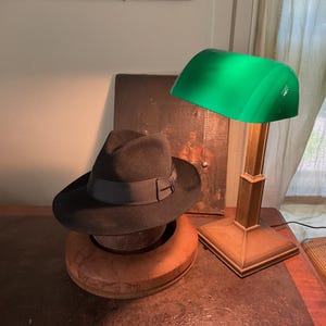 Może przedstawiać: Ciemnoszary kapelusz typu fedora z dopasowaną wstążką, spoczywający na drewnianej formie, obok mosiężnej lampy bankierskiej z jaskrawozielonym szklanym kloszem i łańcuszkiem, z widocznym w tle obrazem w ramie.
