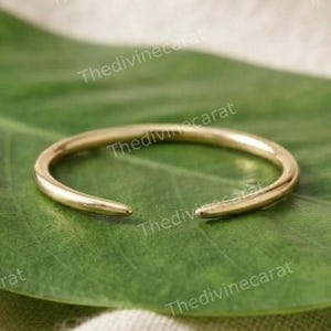 May include: A gold-toned open ring featuring a smooth, polished finish and a slender, circular band with two tapered ends that do not meet, creating a small gap in the centre of the design, showcasing a simple and clean aesthetic.