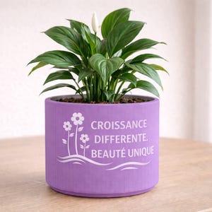 Peut inclure: Un pot de fleurs cylindrique violet vif contenant un lys de la paix vert avec une fleur blanche. Le pot présente un motif blanc de deux fleurs et le texte CROISSANCE DIFFERENTE. BEAUTÉ UNIQUE imprimé sur la surface avant.