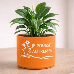 Peut inclure: Un pot de fleurs cylindrique orange vif orné d'un motif blanc représentant deux fleurs et le texte "JE POUSSE AUTREMENT". Une plante de type lys de la paix aux feuilles vertes et dotée d'une unique fleur blanche est plantée à l'intérieur.