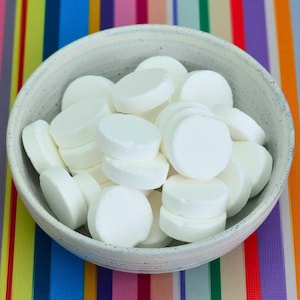 May include: A speckled, off-white ceramic bowl filled with a collection of smooth, opaque white circular tablets. The bowl has a slightly textured, matte finish and a rounded shape, containing numerous stacked and overlapping round white discs.