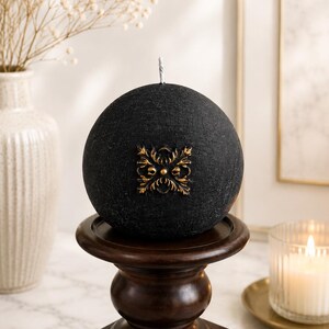 May include: A spherical black candle with a textured surface and a centered gold-colored metallic emblem featuring an ornate floral design. The candle has a single white wick at the top and is positioned on a dark brown wooden pedestal stand.
